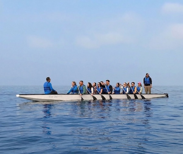 Sakin ve açık denizde beyaz renkli uzun bir dragon bot üzerinde kürek çeken, mavi can yeleği giyen yaklaşık 15 kişilik bir grup. Teknenin ön kısmında yönlendirici konumunda oturan bir kişi, arka kısmında ise ayakta duran bir rehber bulunmaktadır. Katılımcılar gülümseyen ve neşeli ifadelerle pozisyonlarında durmaktadır. Arka planda açık mavi gökyüzü ve sakin deniz görülmektedir.