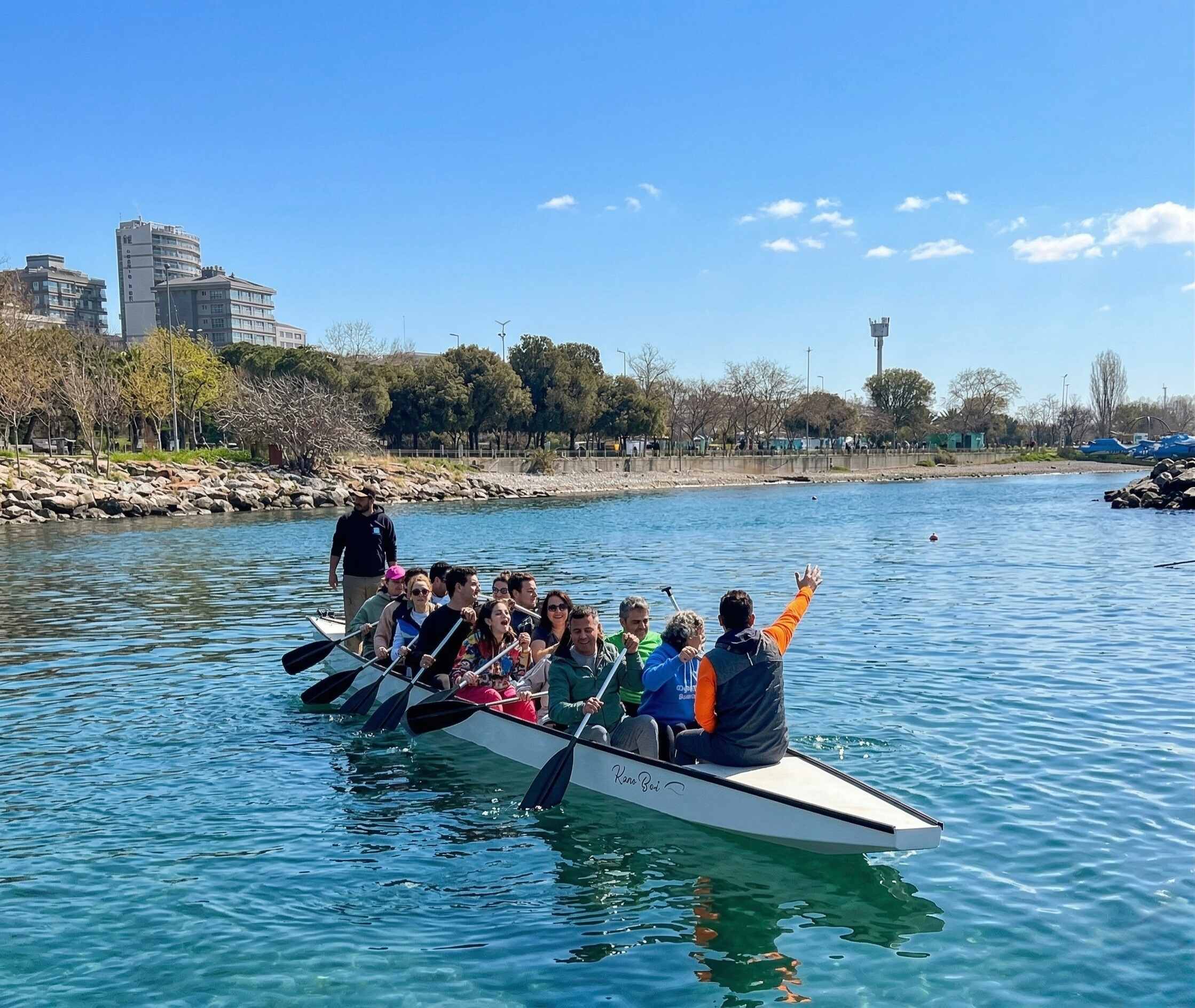Güneşli bir günde turkuaz renkli kanalda beyaz dragon bot ile kürek çeken neşeli bir grup. Teknenin ön kısmında bir katılımcı elini havaya kaldırarak sevinç ifadesi göstermekte, diğerleri gülümseyerek kürek çekmektedir. Teknenin arkasında ayakta duran bir rehber bulunmaktadır. Arka planda taş rıhtım, ağaçlık sahil parkı ve apartman binaları görülmektedir.